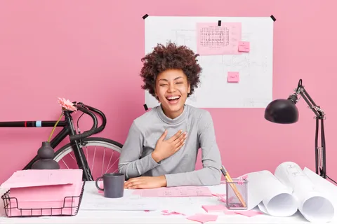An individual laughing while sitting at a desk in a pink room