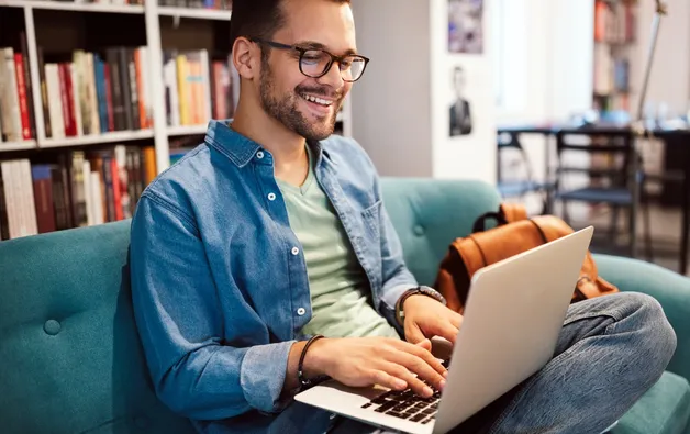 A smiling student working on laptop, sitting on a couch